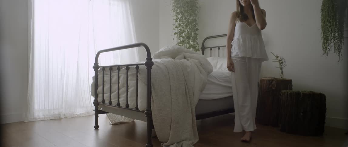 Woman sits at the edge of a bed and gets up to open the curtains