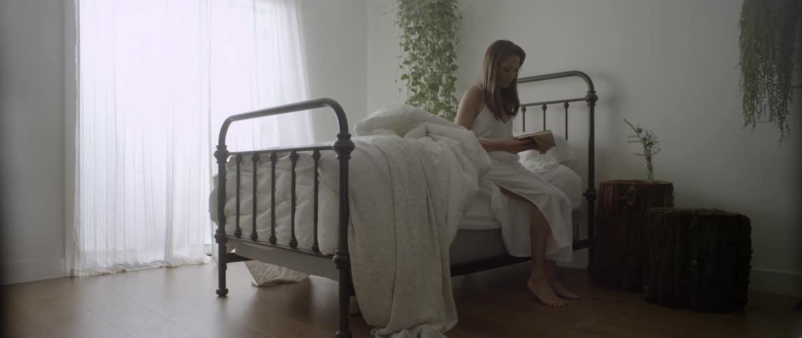 A woman is sitting on the edge of her bed and opening up her journal