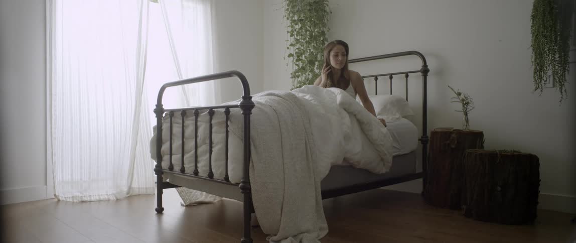 A woman sits up in bed as the curtains blow in the breeze