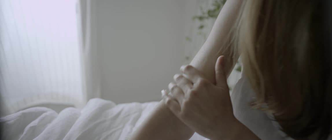 A woman smiles as she strokes her arm while sitting on a bed