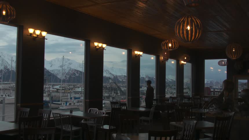 A man stands in a restaurant looking out the window to some mountains and boats