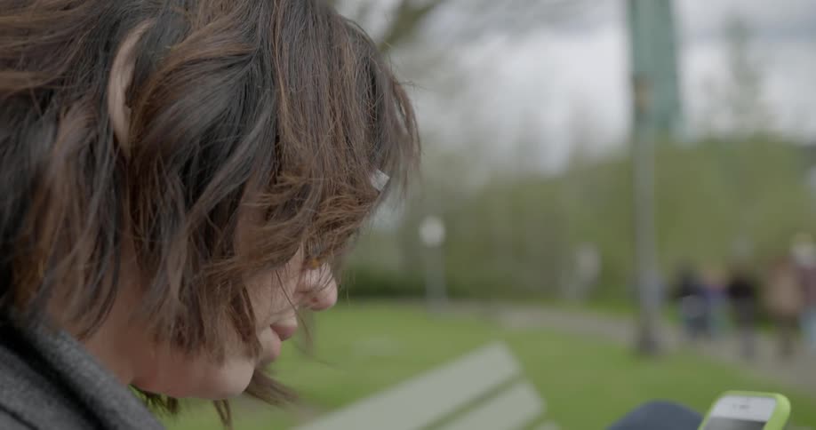 A young woman looks at her cell phone as she sits on a park bench