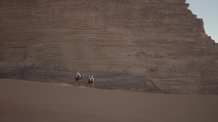 Two camels travel across the desert in front of a mountain