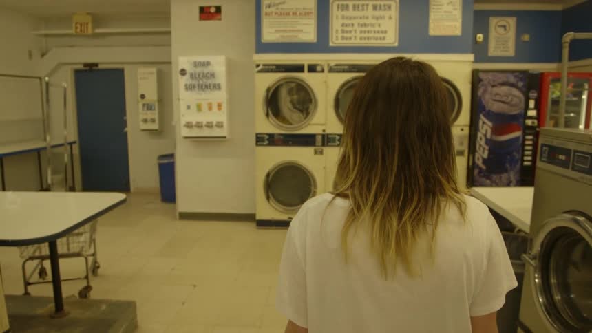 A blond woman walks toward a washer in a laundromat