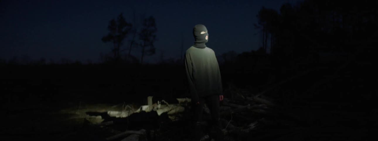 A young man with a ski mask on is standing in the woods while a strobe light is flashing