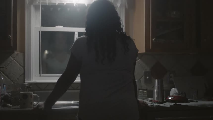 A woman leans on a kitchen sink as she stares out a window at night