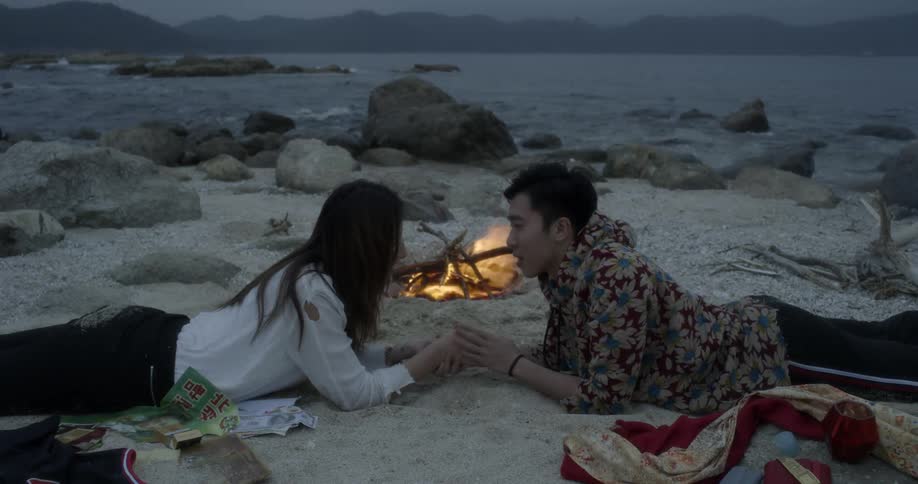 A man and woman lay on the sand as a campfire burns
