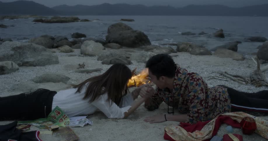Man and woman arm wrestle in front of a small fire on the beach