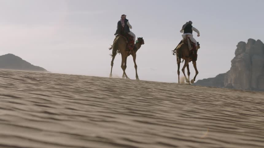Two men ride camels over the sand