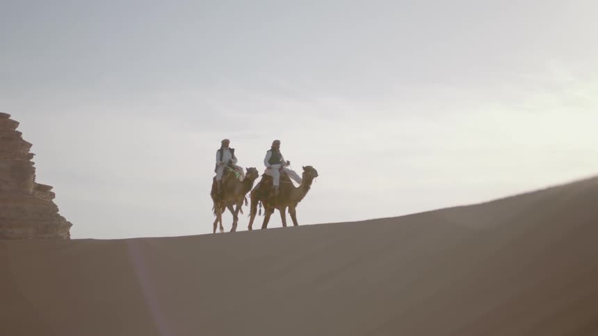 Two men ride camels across a desert hill