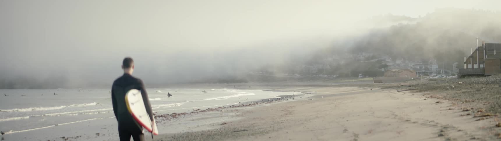 A surfer walks on a beach as fog hovers