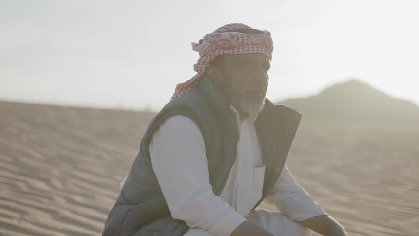 A man wearing a vest and white shirt sits in the desert