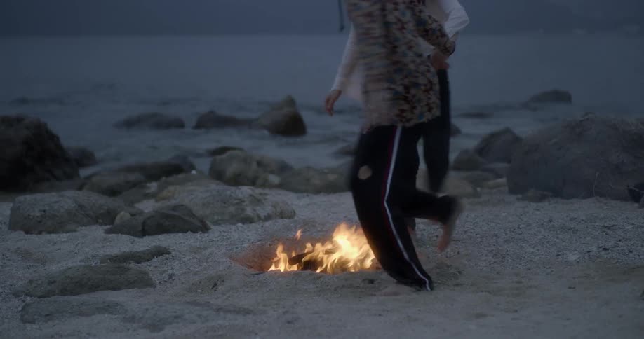 A couple dance around a campfire on a beach at dusk