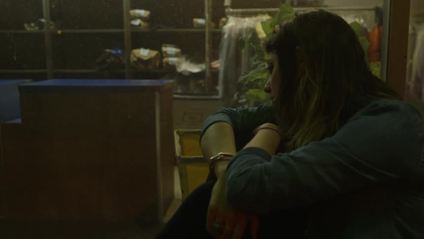 A woman hugs her knees near a window
