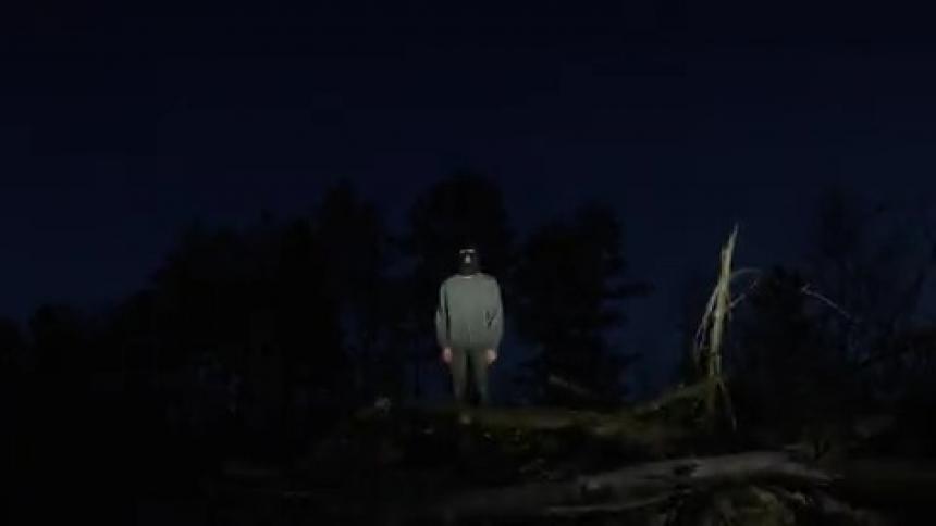 A man with a face mask is standing in the middle of the woods at night with a strobe light on him