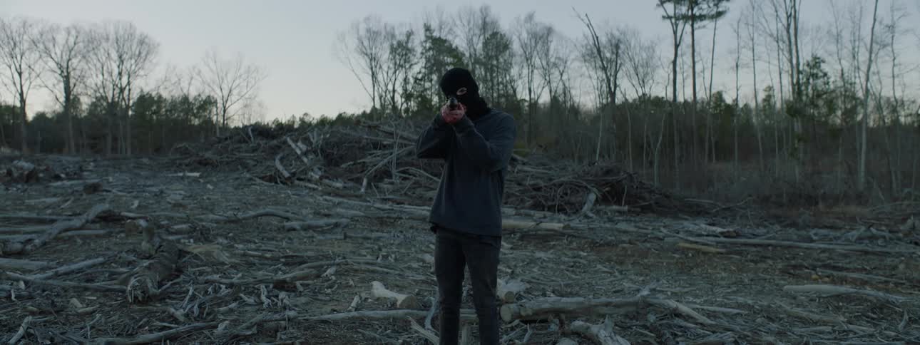 A man with a ski mask points a rifle in the woods