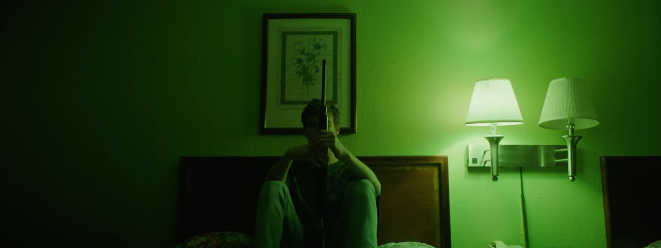 A man is holding a rifle and sitting on the bed in a hotel room