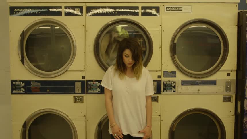 Woman at a laundromat stands in front of dryers looking at the floor