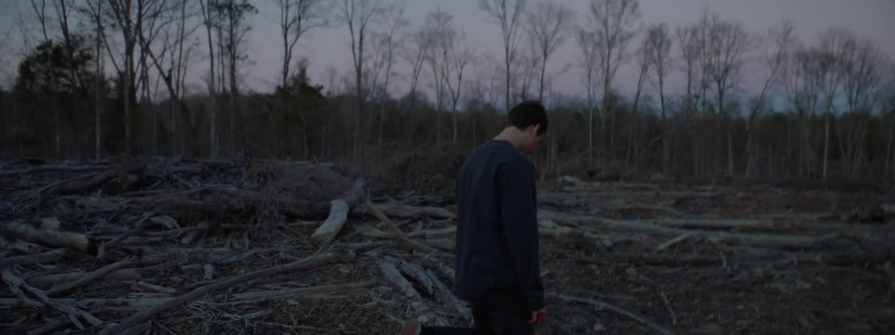 A young man with blood on his hands is kneeling on the ground in a forest
