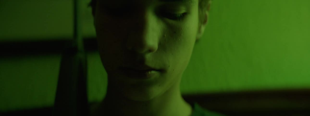 A young man is sitting in a green lit room with a light flashing while holding a gun