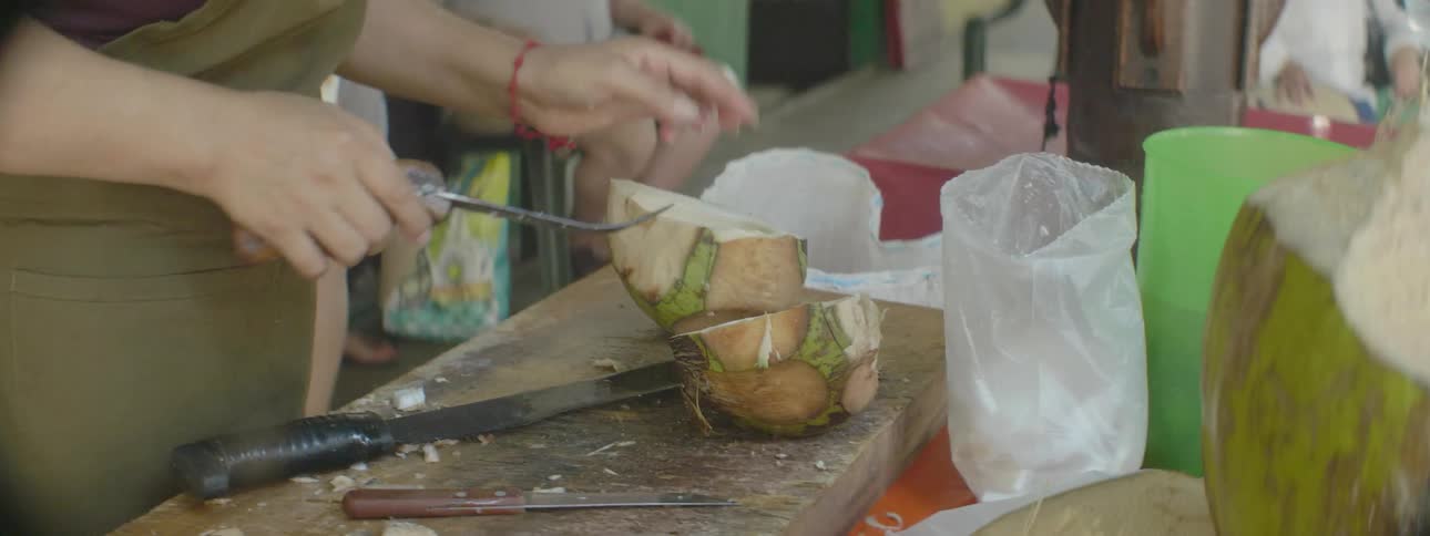 A street food vendor is splitting a coconut and scooping out the inside into a bag for a customer