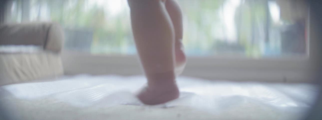 A baby in a living room walks to a cushion and plops down