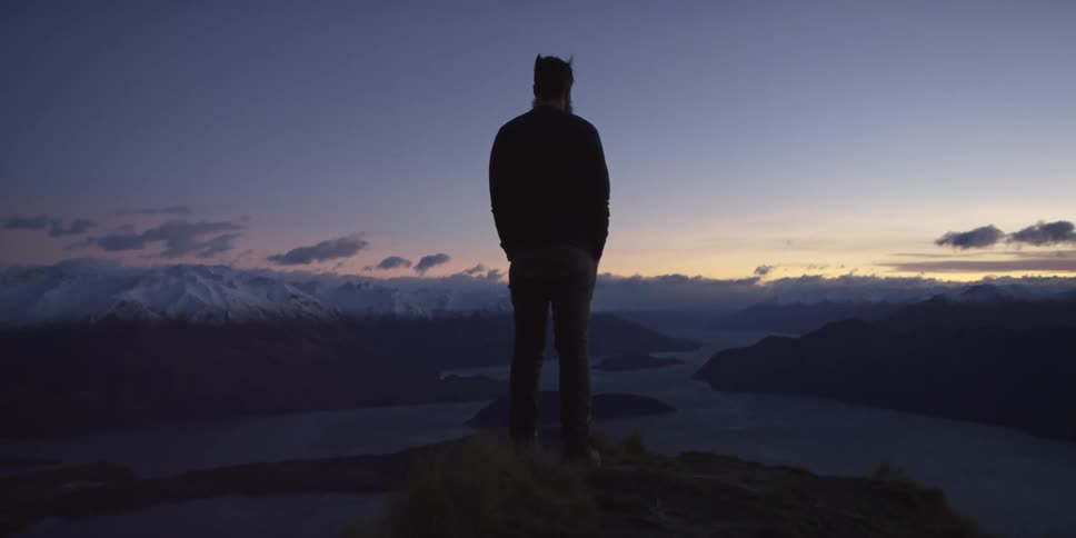 A man stands on a peak overlooking a river at dusk