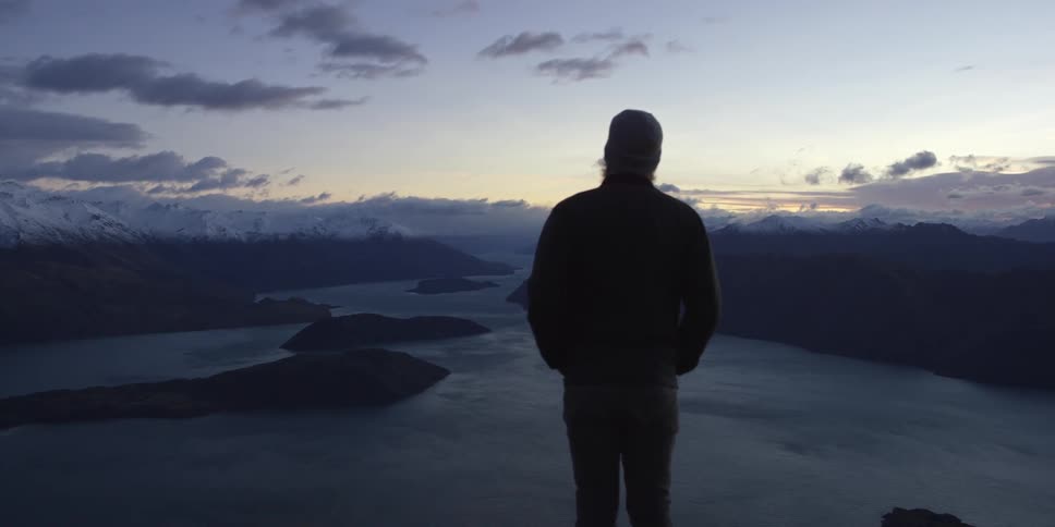 A person stands at the edge of a point looking out over a body of water surrounded by mountains