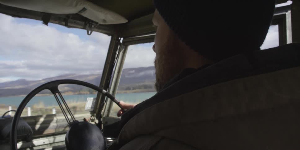 Bearded man driving an older truck past a river