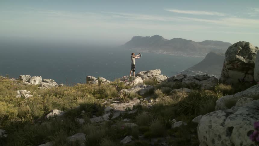 Man holds his arms out standing at the edge of a rocky cliff by water