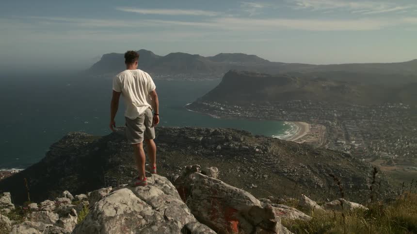 A man is standing on the edge of a cliff overlooking the ocean and a small town