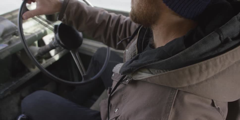 A white man with a beard drives a vehicle down a street