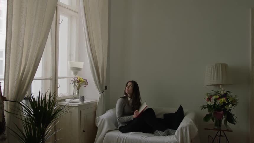 Woman reading a book indoors takes a look up and out a window behind her
