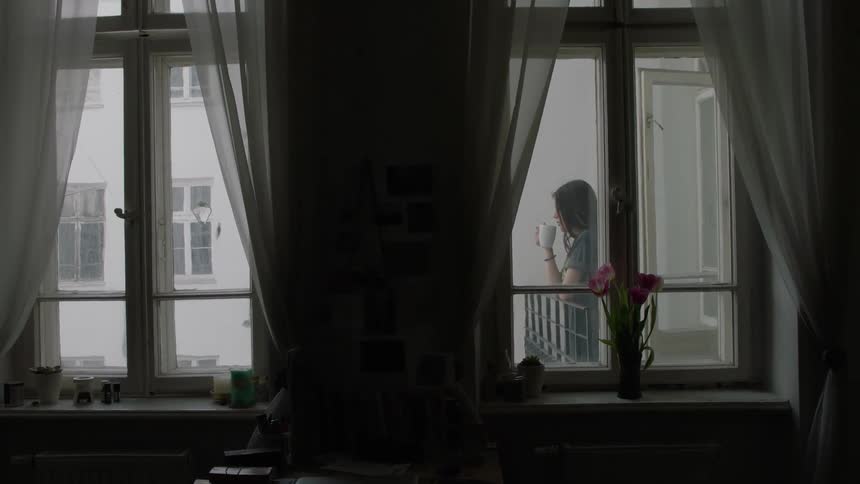 A woman drinks from a mug as she stands on a balcony
