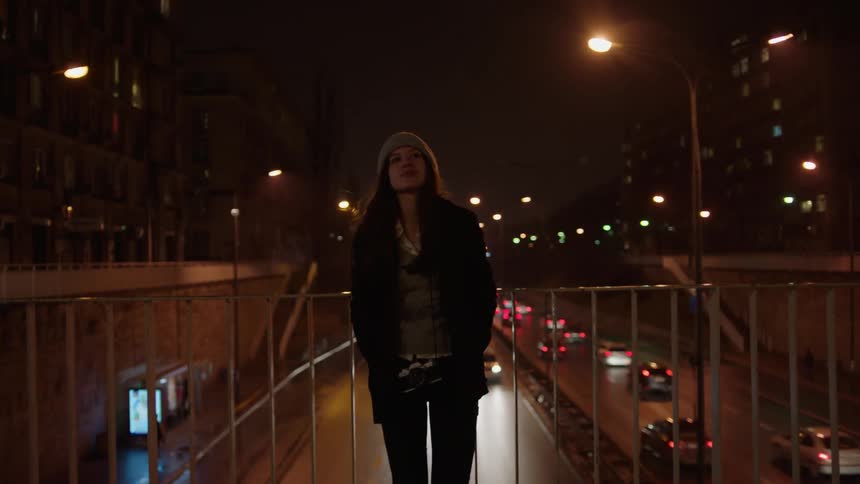 A woman stands on a city overpass at night