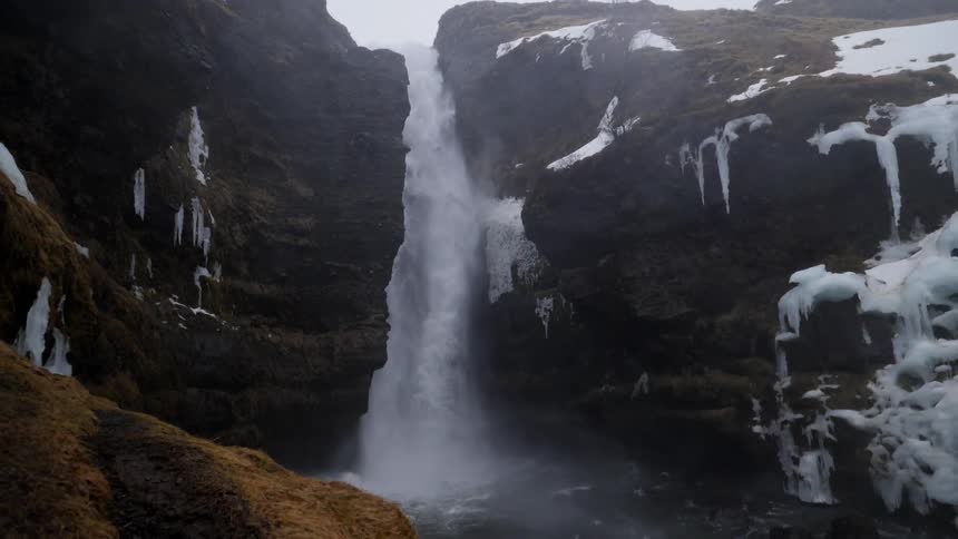 Tall waterfall flowing over edge of rock cliff with patches of snow and ice