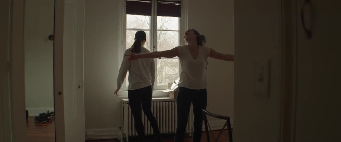 Two women indoors as one looks at the window another spreads her arms and spins around once