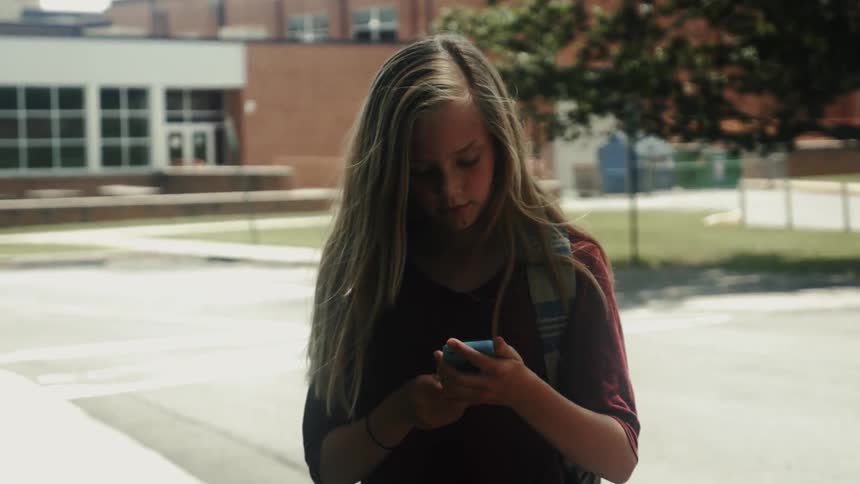A girl standing outside pulls out her cellphone and looks at it