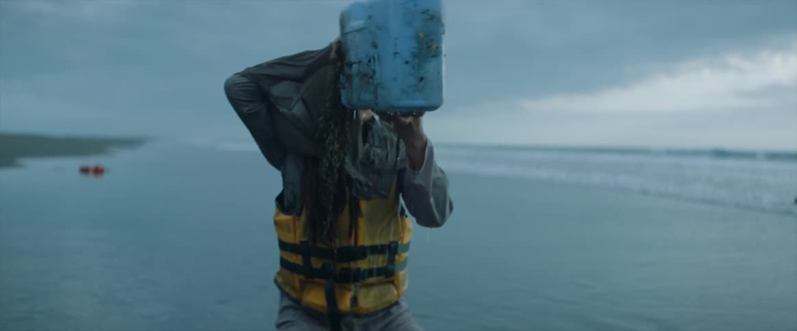 A woman crawls out of the ocean and drinks from a washed up container