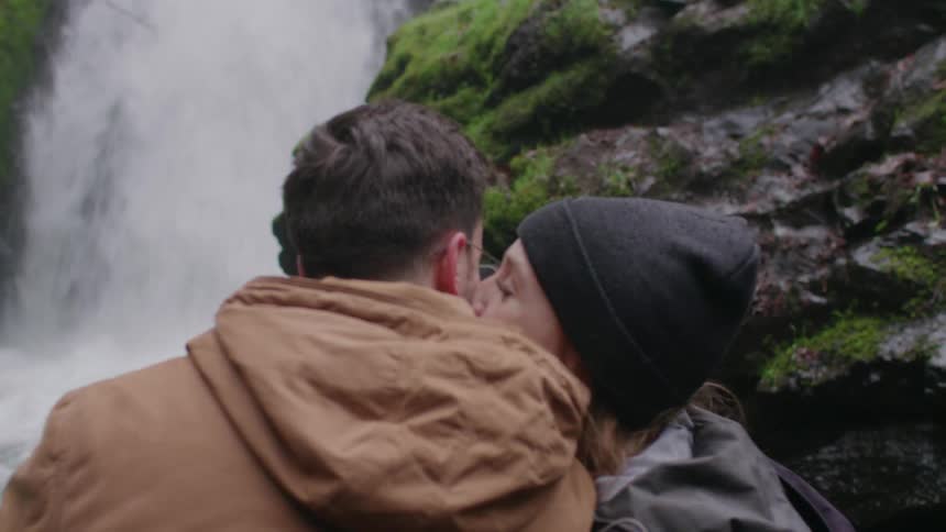 A man and woman kiss while standing next to a waterfall