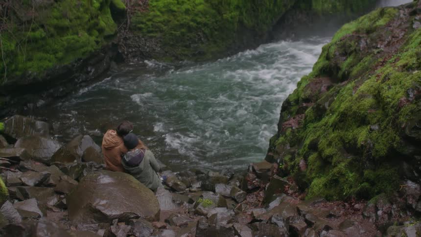 Two people sit on rocks by a flowing river