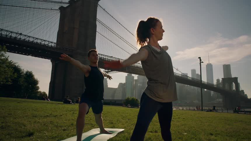 Two women do yoga by a bridge