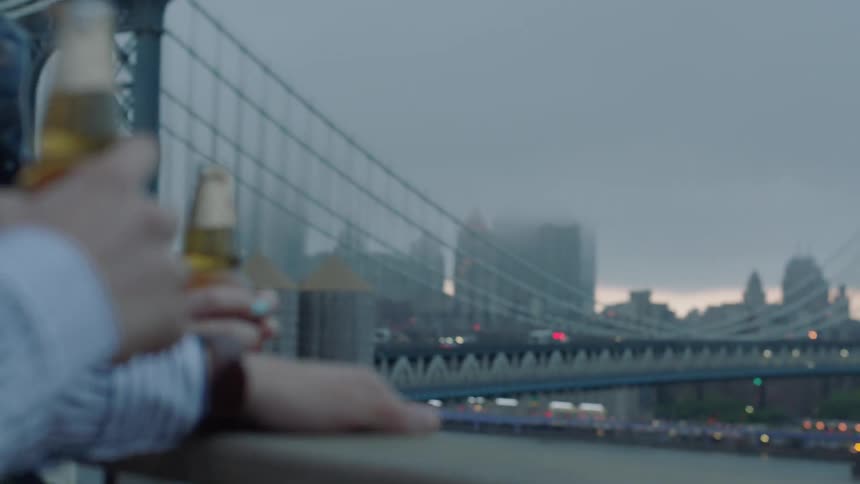 Two people drink beer outside by a bridge