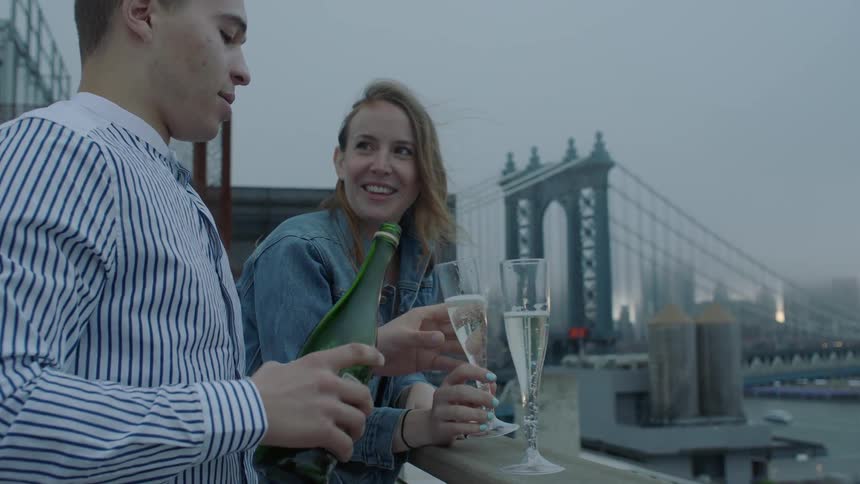 Man pours champagne for himself and a woman on a balcony overlooking a city