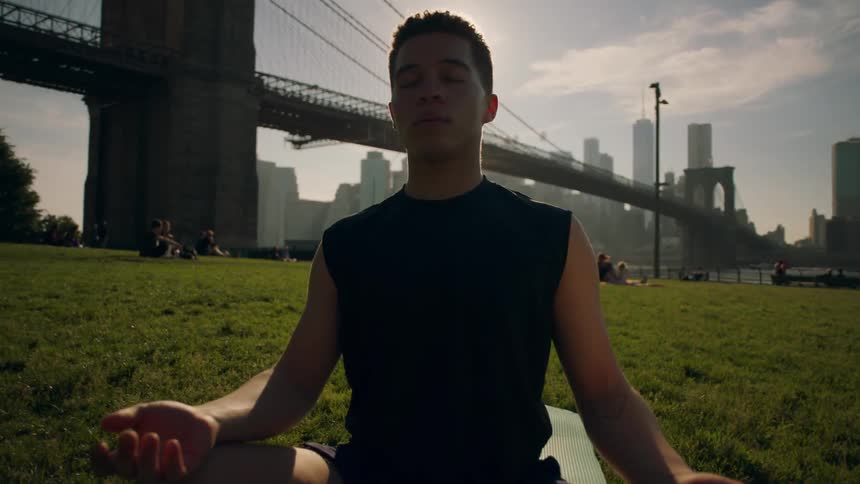 Man sits meditating on a grassy field near a bridge