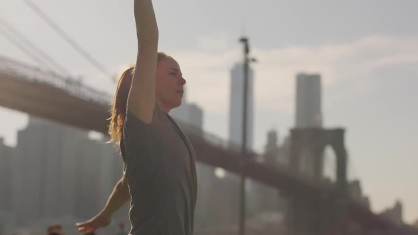 A woman stretches in a city park near a bridge