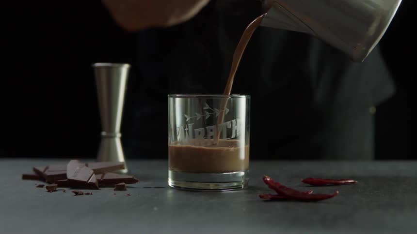 Person pours liquid into a glass on a table with chocolate and chili peppers by it