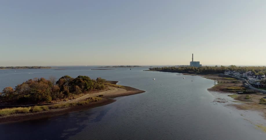 A small island is being filmed by a drone with a larger body of land and lighthouse is in the background