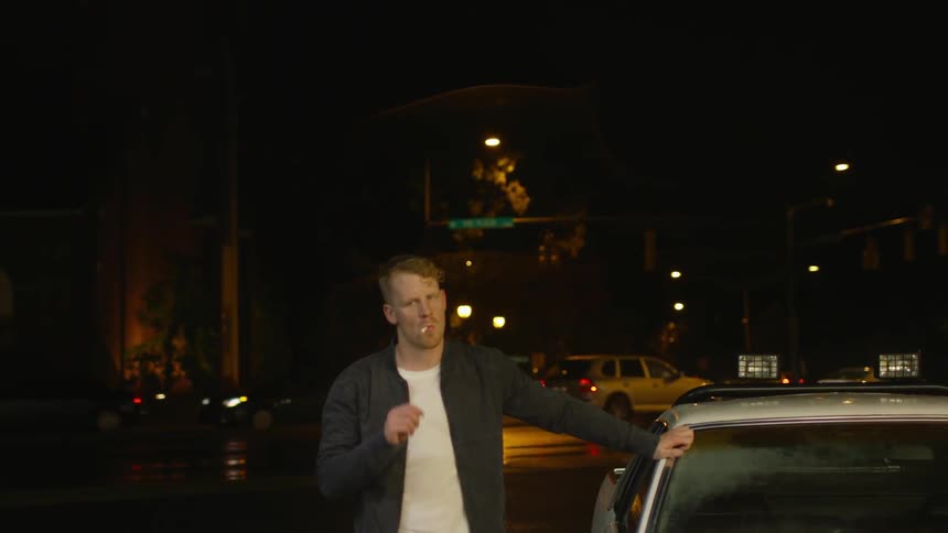 A man stands with his hand on a car while smoking a cigarette