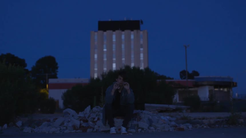 A teenage male sits on a rock pile crumbling leaves at dusk